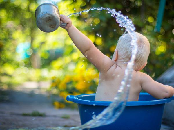 Ein Kind in einer Plastikwanne. Es schüttet Wasser mit einem Becher mit großem Schwung heraus.