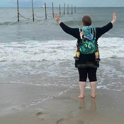 Ein Foto von Stephanie, die am Strand mit den Füßen im Meer steht. Ihren Sohn trägt sie in der Manduca auf dem Rücken.