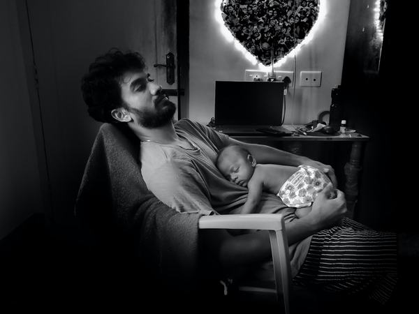 Aufnahme eines sitzenden Mannes mit einem schlafenden Baby auf ihm liegend.