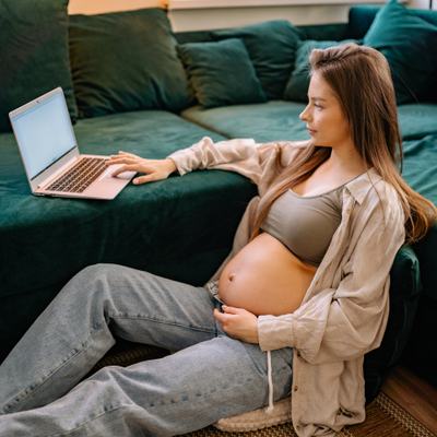 Eine schwangere Frau sitzt auf dem Boden neben dem Sofa und schaut in ein Notebook.