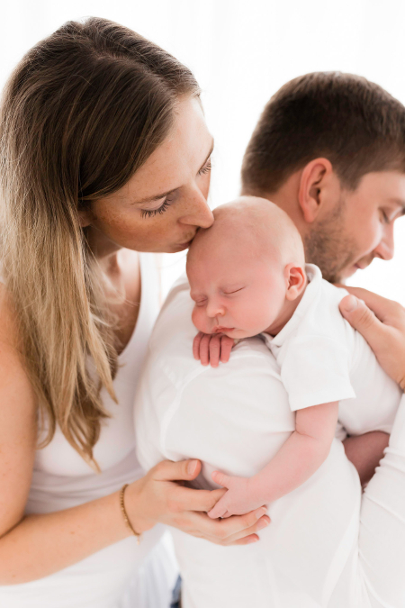 Familienfoto. Der Papa hält das Baby auf dem Arm während die Mama die Hand hält und den Kopf küsst.