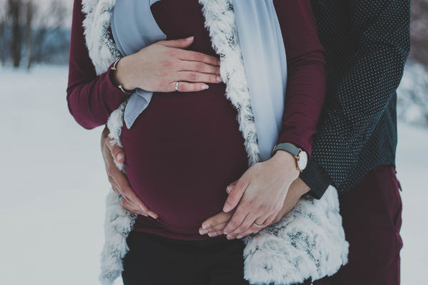 Eine schwangere Frau wird von ihrem Partner umarmt. Er steht hinter ihr und hält ihren Bauch.