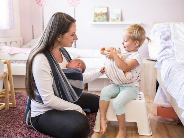 Eine Frau trägt ein Baby im Tragetuch während sie mit einem Kleinkind am Boden spielt.