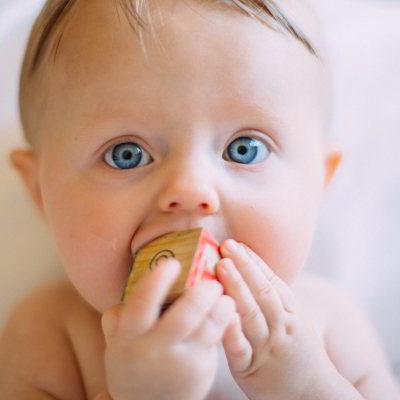 Ein Baby mit hellblauen Augen. In seinen Händen hält es einen Würfel, den es sich in den Mund steckt, um ihn zu erkunden.