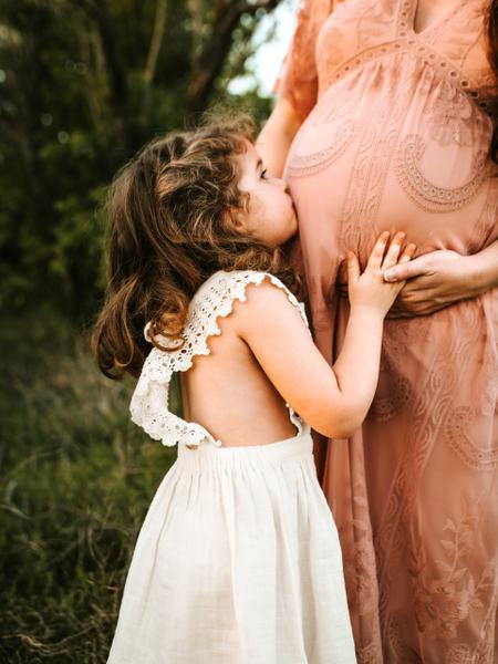 Detailaufnahme einer Frau mit Babybauch. Daneben steht ein Kind und küsst den Bauch.
