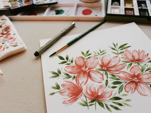 Aufnahme eines Tisches, auf dem ein Blatt Papier mit gemalten Blumen, sowie die Malutensilien liegen.