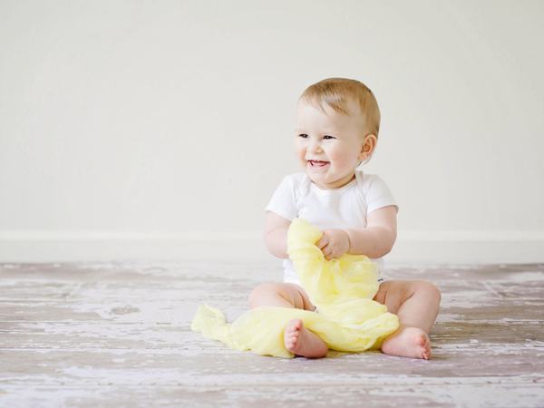 Ein Kleinkind sitzt lächelnd auf dem Boden. Es hat etwas scheinbar plüschiges in der Hand.