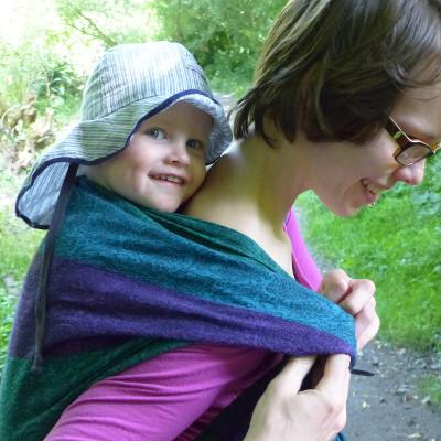 Ich trage meinen Sohn im Tragetuch. Das Tragetuch ist ein grün, violett gestreiftes Tuch von Ellevill. Noah trägt einen Sonnenhut.