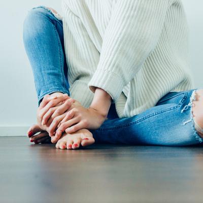 Eine junge Frau mit zerrissener Jeans sitzt auf dem Fußboden. Sie trägt einen weißen Sweater und hat rot lackirte Nägel. Sie ist dabei barfuß.