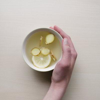 Eine Hand hält eine Tasse Tee fest. In der Tasse schwimmen Ingwer- und Zitronnenstücke.